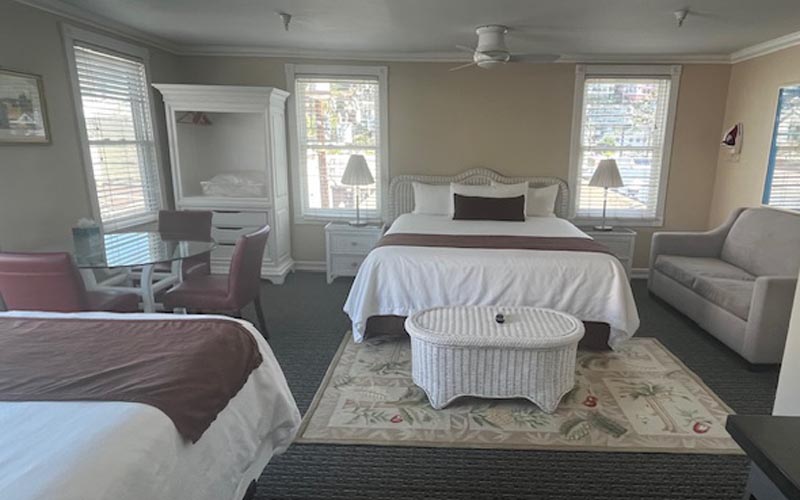 Spacious hotel suite with two white beds, a matching wicker coffee table, a beige sofa, and a glass dining table by large windows.