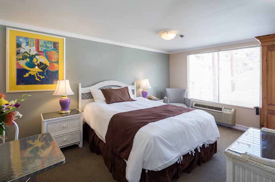 Hotel bedroom with a white bed and brown accents, two wicker nightstands with purple lamps, and a colorful framed painting on a gray wall.